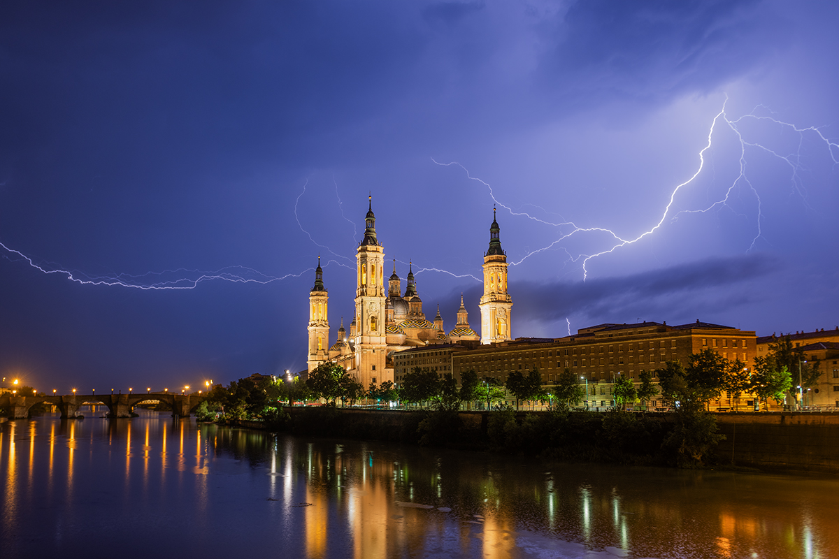 Esta fotografía es el resultado de la constancia. En un viaje a la comunidad de Aragón, decidí intentar captar una tormenta con la preciosa basílica del Pilar en Zaragoza debido a la previsión de tormentas nocturnas en la zona. Cuando vi que finalmente la tormenta llegaba tuve que buscar una buena localización orientada a la tormenta con la basílica en primer plano. No fue una tarea fácil, era fin de semana y había a esas horas mucha gente de fiesta por la ciudad por lo que lo primero era aparcar el coche, cuando lo conseguí cargado con trípode, cámaras paraguas al puente sobre el que tomé la fotografía y a esperar hasta que un rayo me cruzó la imagen al cabo de bastantes minutos.
