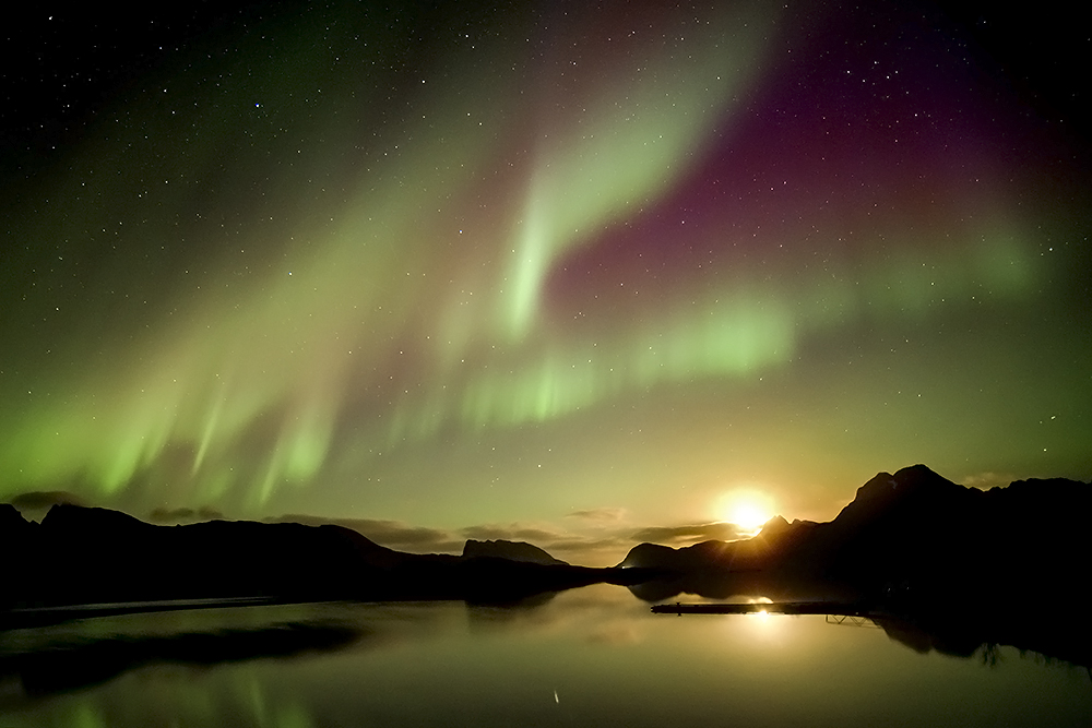 La luna en cuarto creciente discurría muy bajo por el horizonte, mientras que la aurora boreal aparecía sobre el cielo y con el mar abajo reflejando los colores nos ofrecía todo un espectáculo visual en la noche del 9 de noviembre de 2024 en las islas Lofoten en Noruega  
