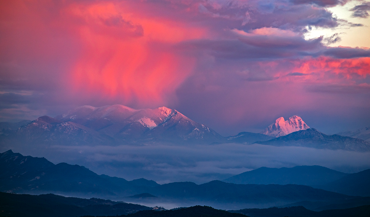 Detrás de los valles cubiertos por nieblas, las primeras luces del sol al amanecer iluminaban una rojiza cortina de nieve movida por el viento sobre la Serra d’Ensija, a la derecha también iluminada por el sol, la emblemática montaña Pedraforca.
Este regalo de la naturaleza lo capturé desde la zona elevada del Santuari dels Munts.

