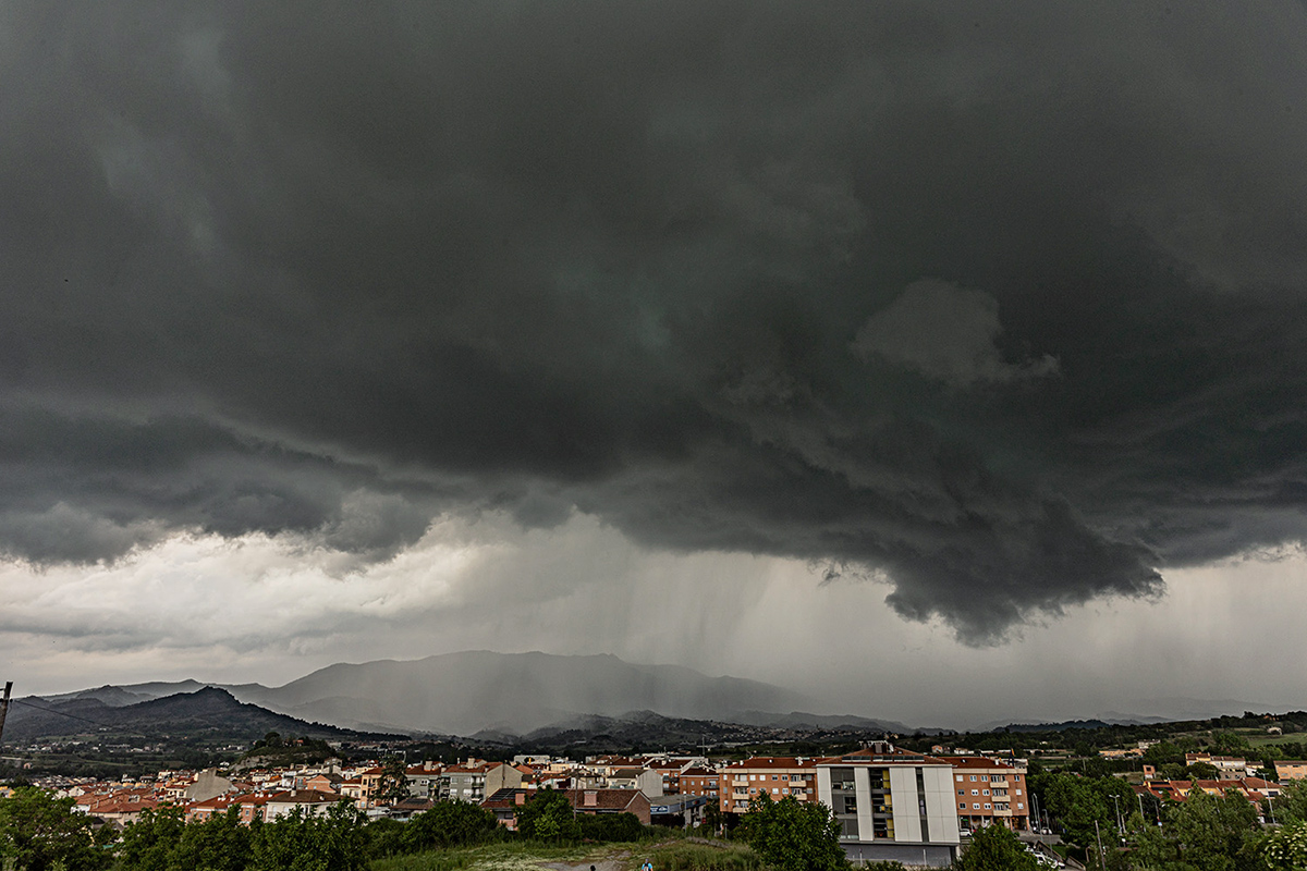 Cielo intensamente cubierto por nubes de tormenta oscuras y amenazadoras, que dominan la escena con una atmósfera inquietante. Las nubes son densas, de tonos gris oscuro y negro. En el horizonte, puede verse una cortina de lluvia intensa cayendo sobre la montaña de Bellmunt, al final de la tarde.

