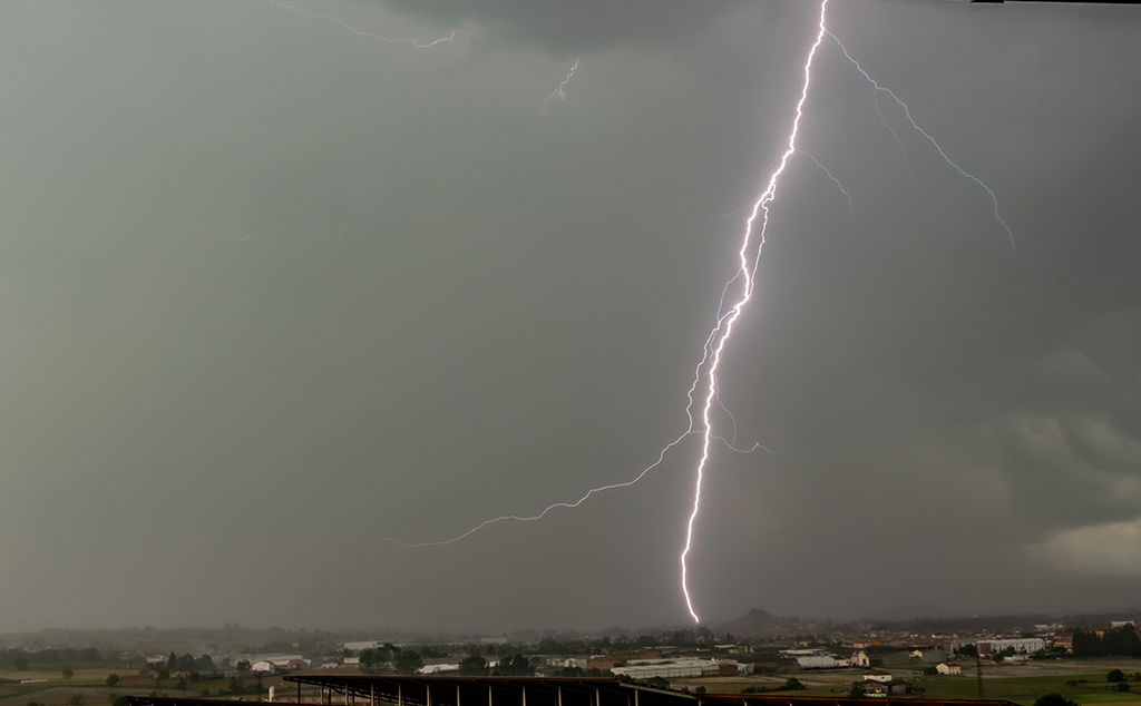 Un potente relámpago impacta sobre el paisaje durante una tormenta diurna. El rayo con su trayectoria irregular y su luz intensa, contrasta de manera espectacular con el fondo gris verdoso.
