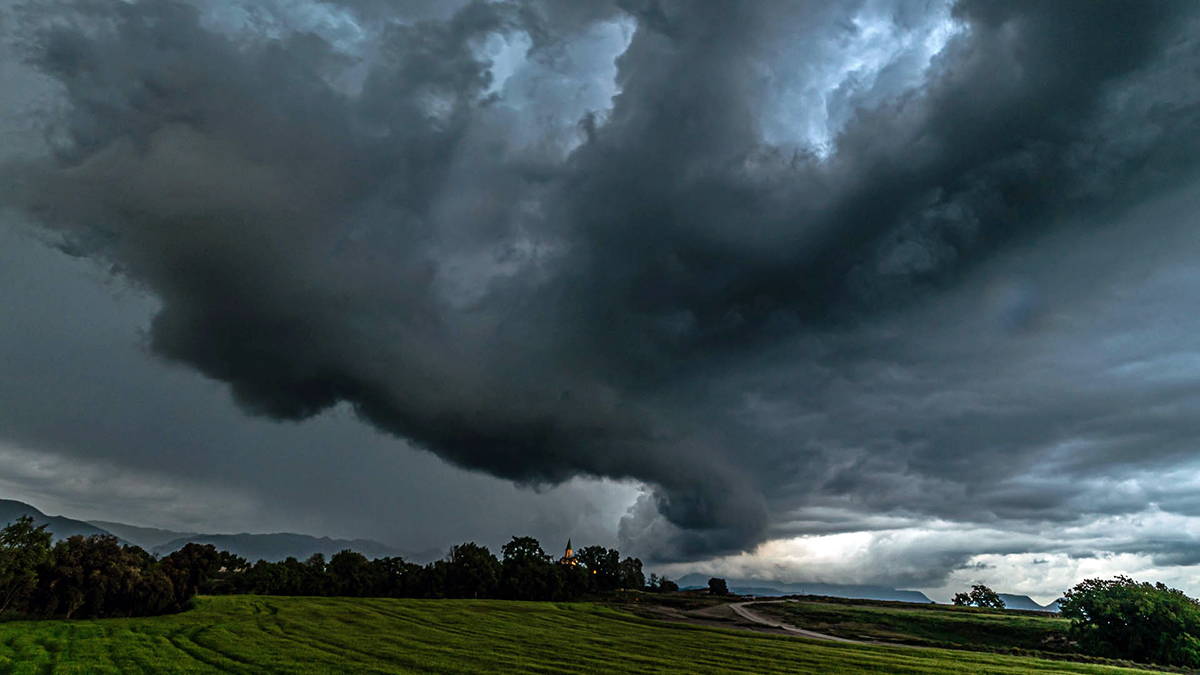  Nube torbellino formándose bajo una enorme masa de nubes oscuras. El cielo está cubierto casi por completo por una estructura nubosa en espiral, densa y turbulenta, que desciende en forma de embudo hacia el suelo, típica de una supercélula o sistema convectivo severo. El paisaje rural se extiende con campos verdes bien definidos y un camino de tierra que serpentea hacia el horizonte. A lo lejos, se vislumbra pequeño el santuario de Puig-Agut iluminado, cuya luz contrasta dramáticamente con la oscuridad del cielo.


