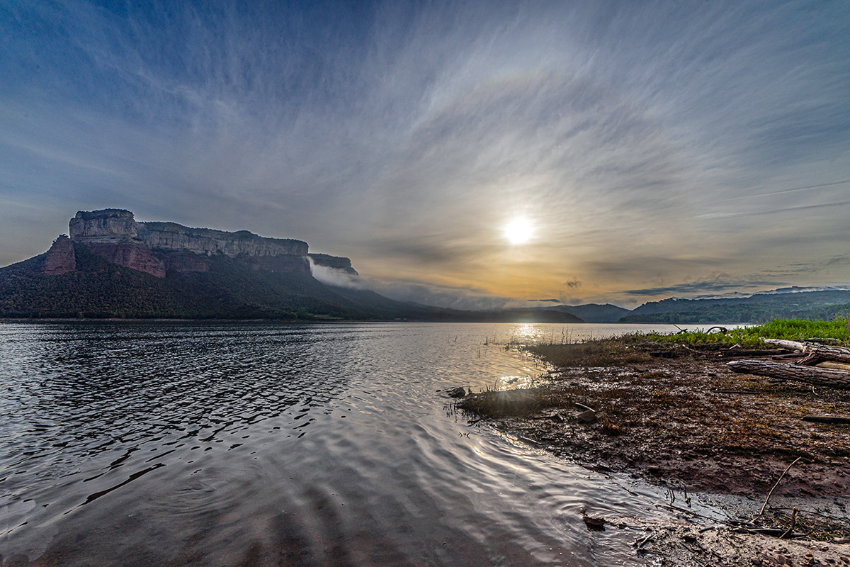 Sereno paisaje del embalse de Sau, acompañado por un fenómeno atmosférico fascinante: un halo solar. El sol, se encuentra parcialmente velado por nubes finas de cristales de hielo, los cuales refractan la luz y crean un círculo luminoso alrededor del astro, dando lugar a este halo casi místico. A la izquierda, se eleva majestuosa la silueta del Puig de la Força, de Tavertet. La luz dorada del sol baña las montañas y las aguas tranquilas del embalse, generando reflejos suaves sobre la superficie ondulada. A la derecha, la orilla fangosa, salpicada de ramas y vegetación baja, ofrece un primer plano contrastante que aporta textura y profundidad a la escena.
