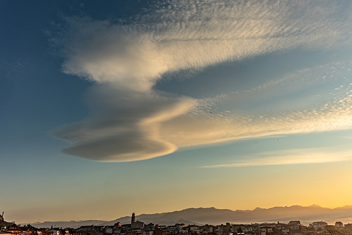 Lenticular al amanecer, capturada al despuntar el día, una majestuosa nube lenticular domina el cielo sobre un paisaje urbano de Sant Hipòlit, aún dormido. Este tipo de nube, con su forma de lente o platillo volador, es el resultado de corrientes de aire húmedo que fluyen a gran velocidad sobre relieves montañosos o irregulares. Al ascender y descender, el aire se enfría y condensa, formando estas estructuras estacionarias y perfectamente contorneadas.
