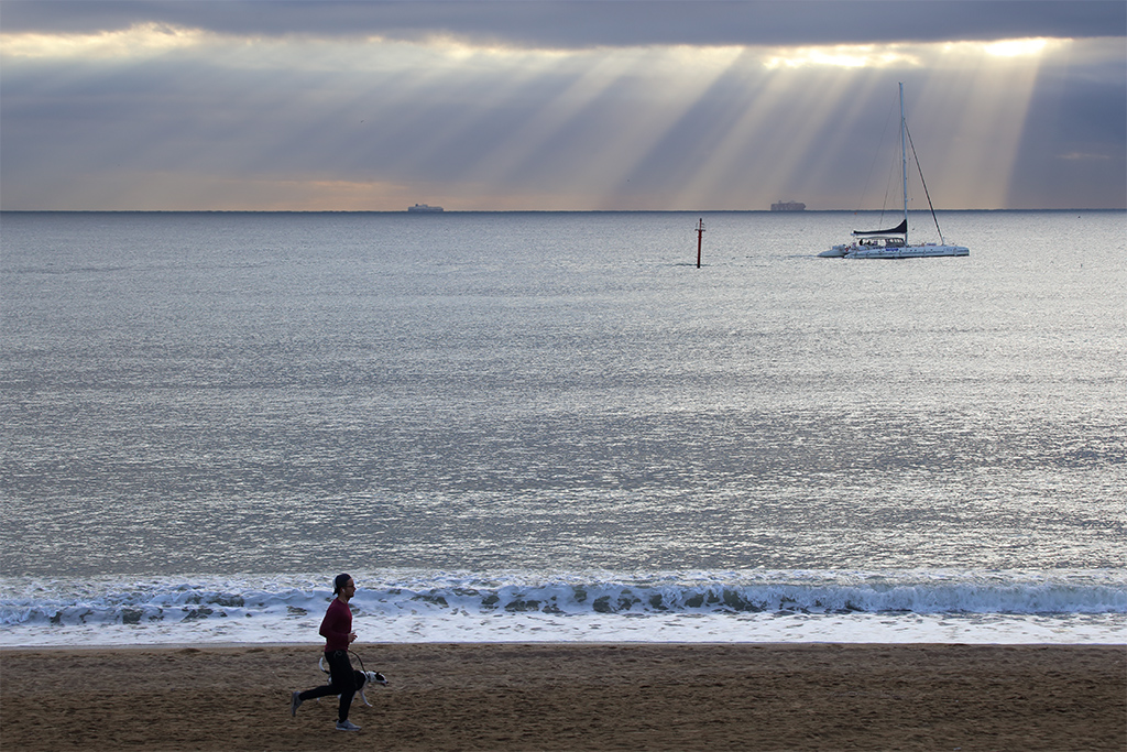 Rayos y barcos
Un chico y su perro corren por la playa de Barcelona mientras por el horizonte se van filtrando los rayos solares entre las espesas nubes que lentamente se van disipando. 
