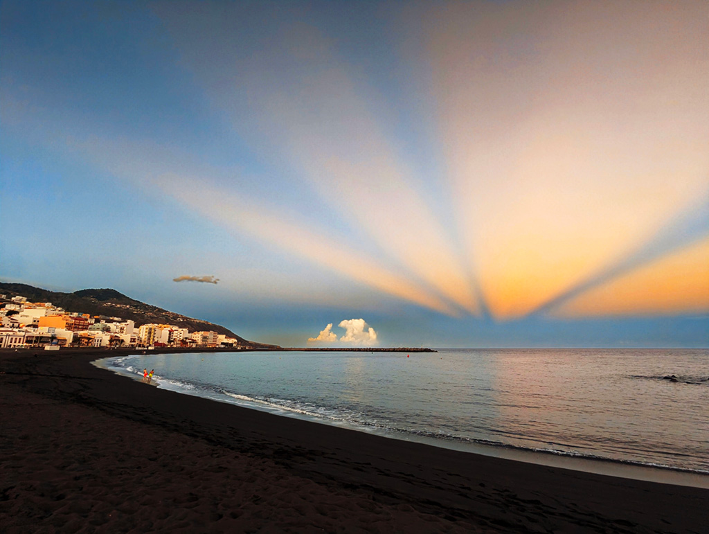 El lunes 18 de noviembre, se capturó una impresionante fotografía de rayos anticrepusculares en la bahía de Santa Cruz de La Palma. Estos fenómenos ópticos son sombras proyectadas por enormes nubes cumulonimbus, formadas en una borrasca fría aislada que se desplazó desde la península ibérica hasta situarse al noroeste de la isla de La Palma. Aunque la borrasca no llegó a impactar directamente en la isla, el servicio meteorológico había emitido alertas por posibles lluvias de hasta 100 litros por metro cuadrado y se decretó una situación de alarma. Sin embargo, la borrasca descargó en el mar y se disipó, dejando como resultado esta espectacular imagen al atardecer del lunes.
Los rayos anticrepusculares son similares a los rayos crepusculares, pero se observan en el lado opuesto al sol en el cielo. Aunque los rayos anticrepusculares parecen converger en un punto opuesto al Sol, la convergencia es en realidad una ilusión. Los rayos son en realidad (casi) paralelos, y la convergencia aparente es al punto de fuga en el infinito. 
La fotografía, tomada con un Pixel 6 Pro sin edición adicional, muestra la intensidad y magnitud de este fenómeno, que duró aproximadamente entre 10 y 15 minutos. Detrás, al oeste de la bahía de Santa Cruz de La Palma, se elevan montañas de casi 2.000 metros de altura, que contribuyen a la formación de estos efectos ópticos.
Las coordenadas geográficas de Santa Cruz de La Palma son:
•	Latitud: 28°41'0.6" N
•	Longitud: 17°45'51.2" O

