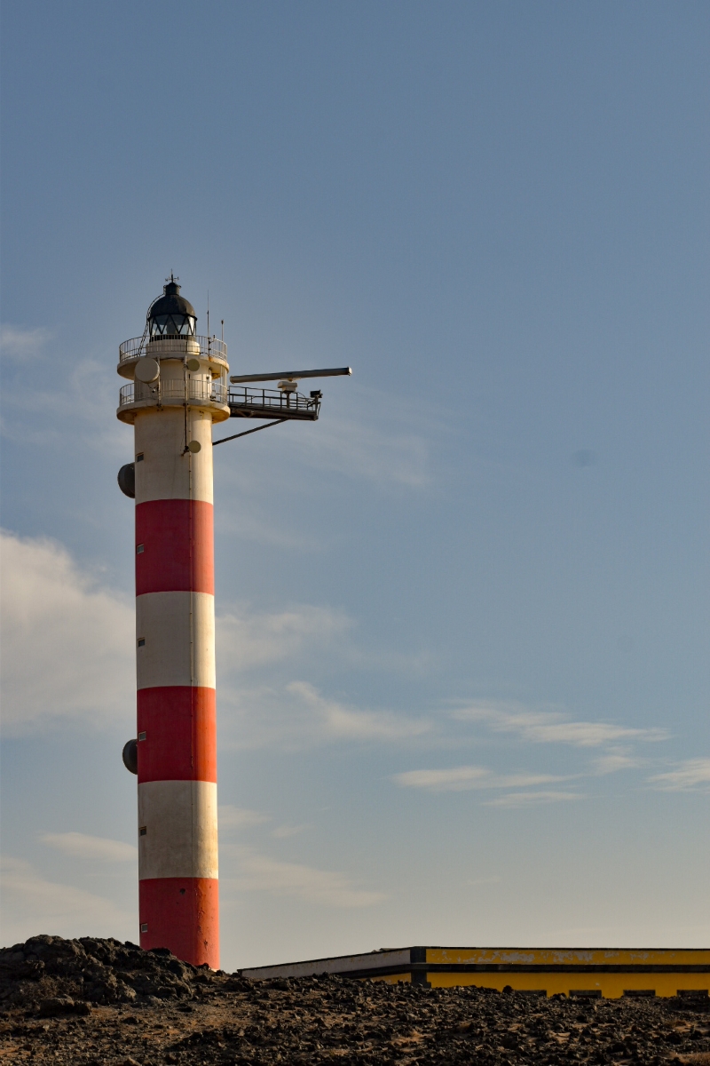 La fotografía muestra el Faro del Porís , en Tenerife, destacando su estructura cilíndrica de franjas rojas y blancas contra un cielo azul despejado. Situado en un paisaje rocoso y árido, el faro representa un símbolo de guía y orientación para los navegantes.
