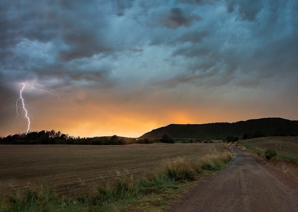 Tormenta al atardecer en Ódena, Igualada (BCN) entre campos de trigo y viñedos donde se puede observar el paso de las nubes de tormenta mezcladas con la luz del sol poniéndose tras las montañas mientras a la misma vez un rayo cielo-tierra hace acto de presencia.
