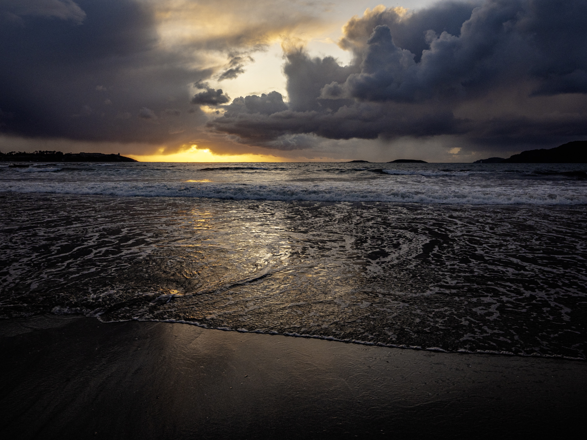 Atardecer primaveral, con lluvia, viento, sol y nubes con un mar en relativa calma que todo lo refleja.
