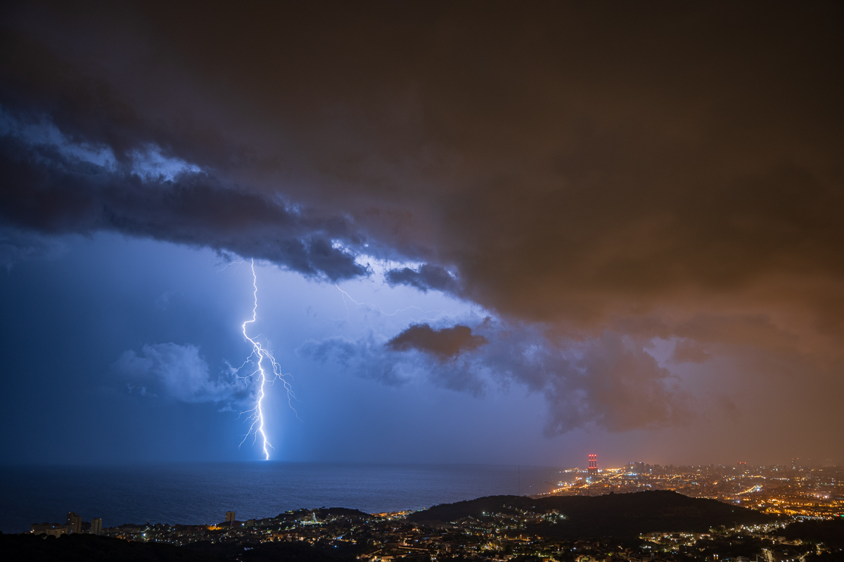 Tormenta en la costa de Barcelona. Otra captura de la noche del 30.8.23, con el rayo impactando en el mar y mostrándonos su enorme tamaño y fuerza. Una combinación perfecta entre colores azules (luz natural) y naranjas (luz artificial).
